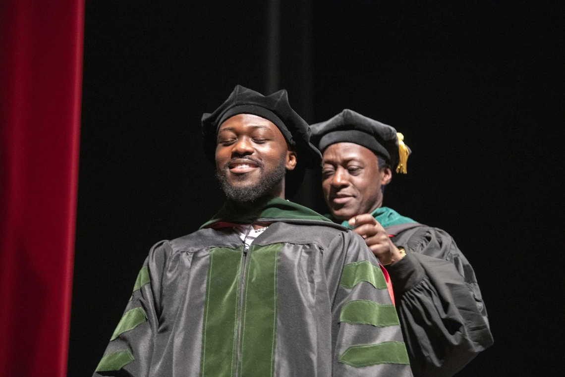 A University of Arizona College of Medicine – Tucson student dressed in a graduation cap and gown has a ceremonial hood placed over his shoulders by his father, who is also dressed in graduation regalia.