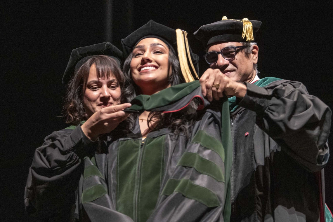 Two University of Arizona College of Medicine – Tucson professors place a ceremonial graduation hood over the shoulders of their daughter. All are dressed in caps and gowns and smiling. 