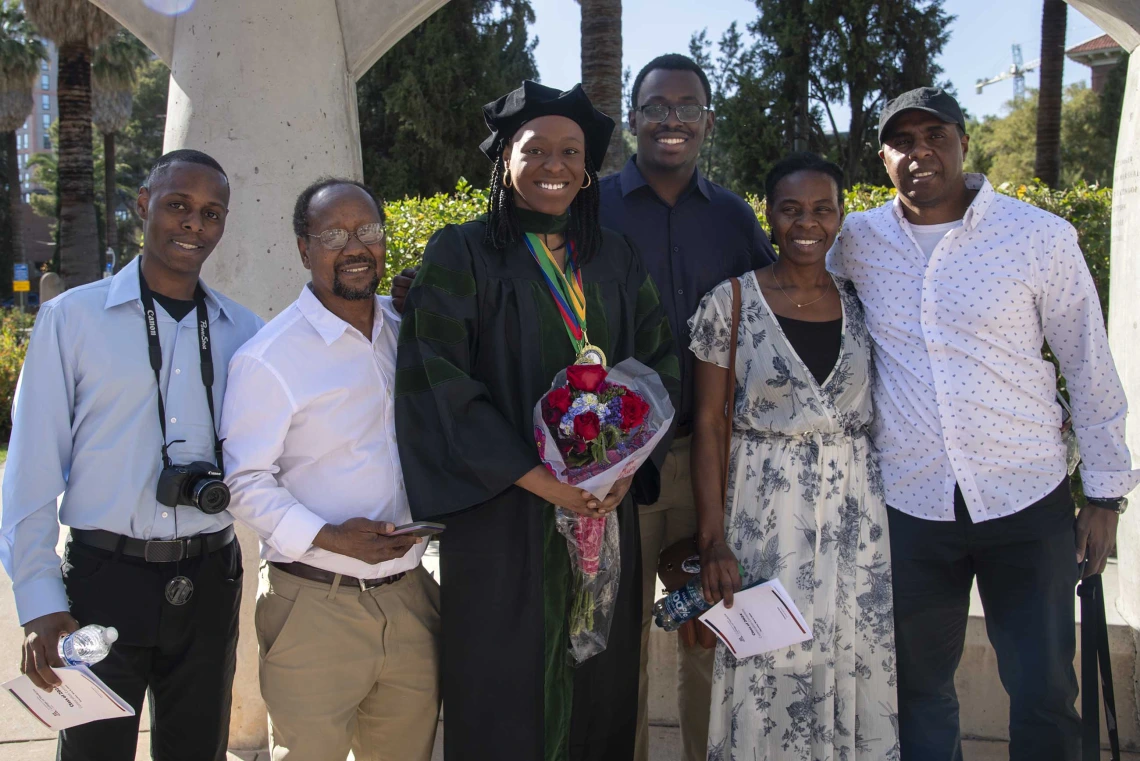 A University of Arizona College of Medicine – Tucson graduate in a cap and gown smiles as she holds flowers while surrounded by family and friends.