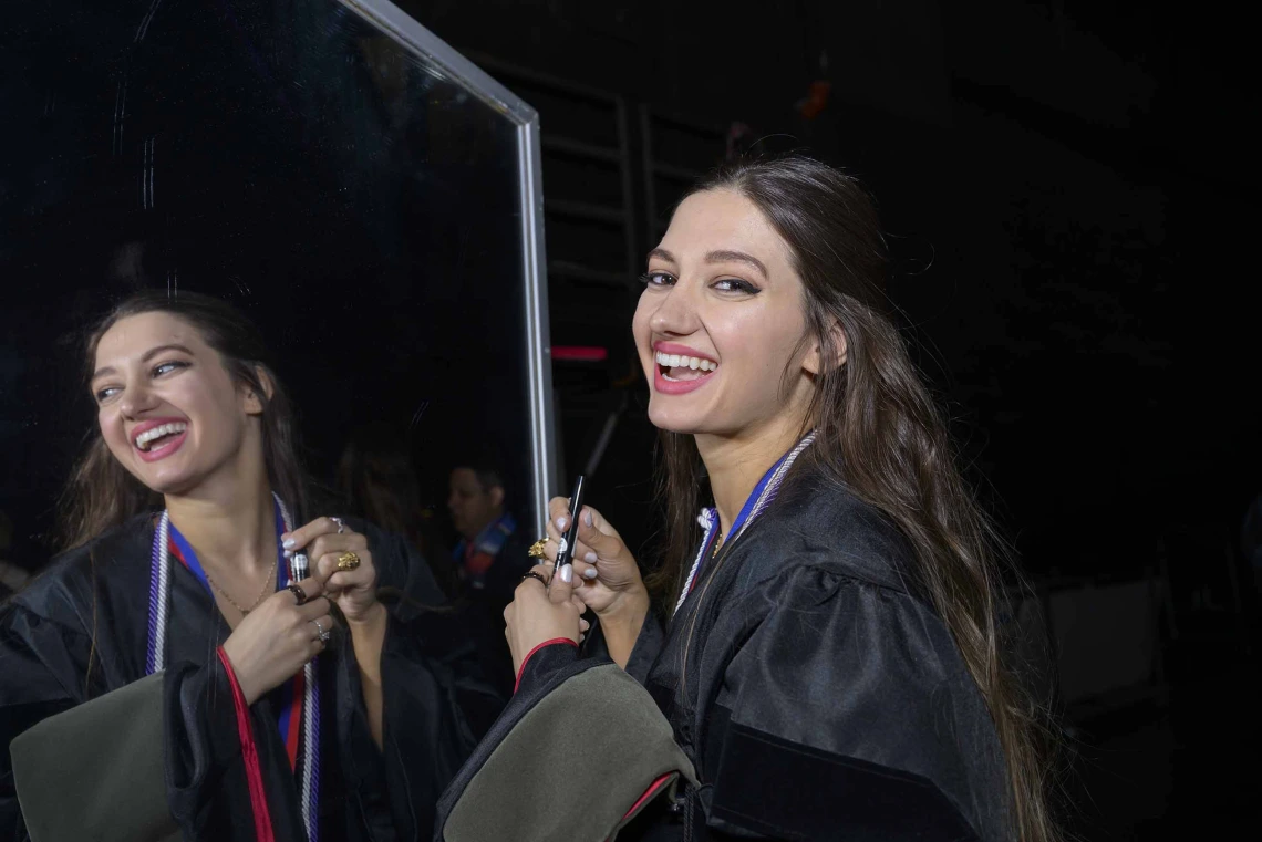 A University of Arizona R. Ken Coit College of Pharmacy student wearing graduation regalia smiles as her image is reflected in a mirror. 