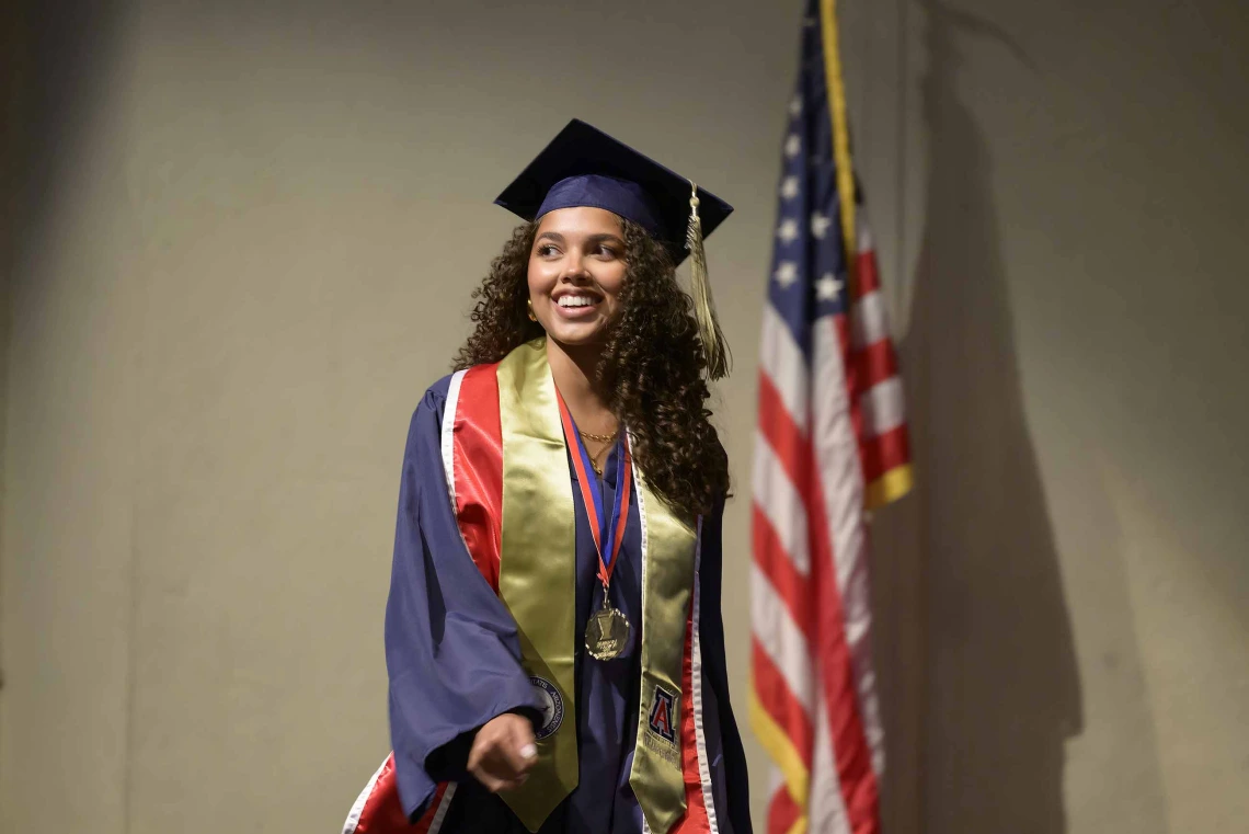 A University of Arizona R. Ken Coit College of Pharmacy student dressed in a graduation cap and gown smiles as she walks in front of an American flag. 