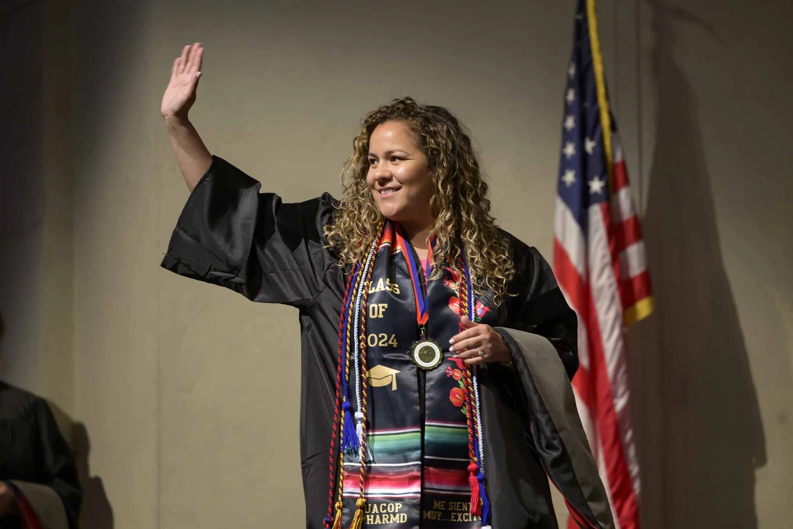 A University of Arizona R. Ken Coit College of Pharmacy student dressed in a graduation gown waves and smiles. There is an American flag behind her. 