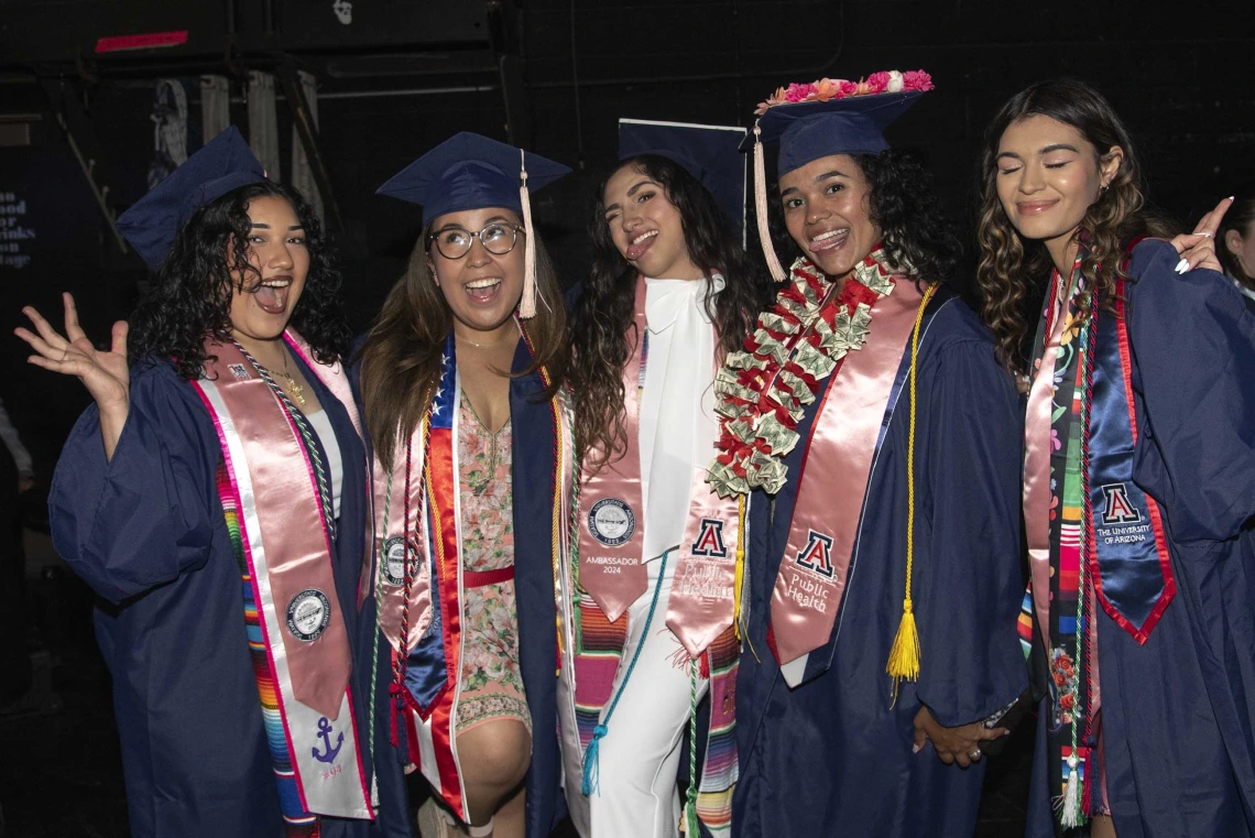 Five University of Arizona Mel and Enid Zuckerman College of Public Health students, all wearing graduation caps and gowns, smile and wave. 