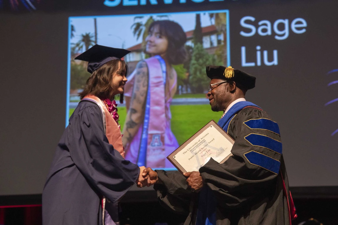 A University of Arizona Mel and Enid Zuckerman College of Public Health student shakes hands with a professor who is about to hand her an award. Both are dressed in graduation regalia. 