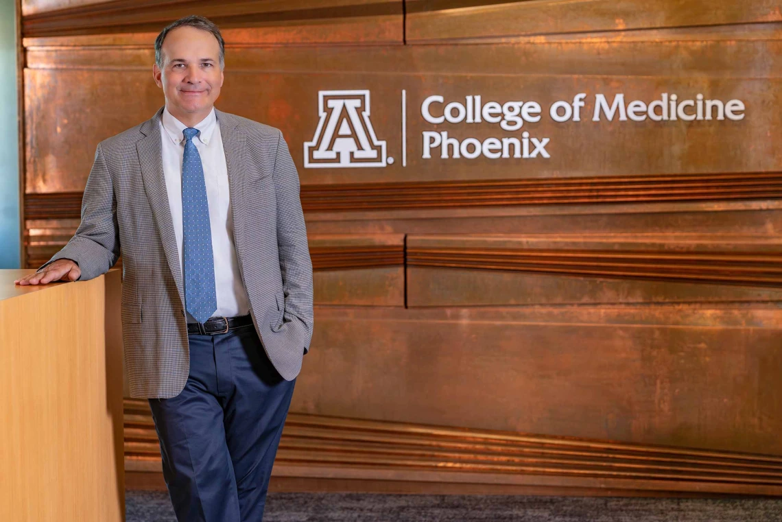 A man in a suit coat stands in front of a wall that has College of Medicine – Phoenix lettering