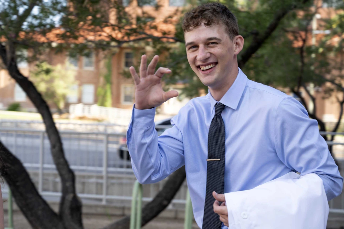 A new University of Arizona College of Medicine – Tucson student flashes the Arizona Wildcats hand sign as he walks outside with a medical white coat draped over his arm. 