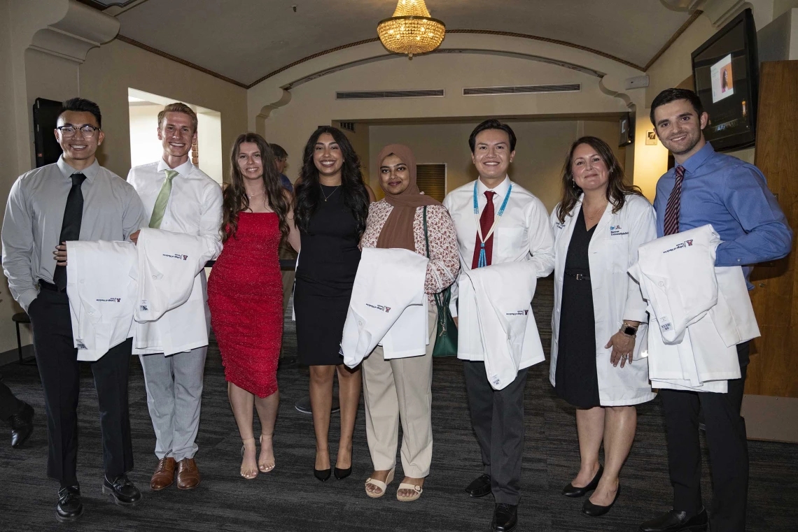 A University of Arizona College of Medicine – Tucson professor wearing a white medical coat stands with seven new medical students, all with white coats draped over their arms.