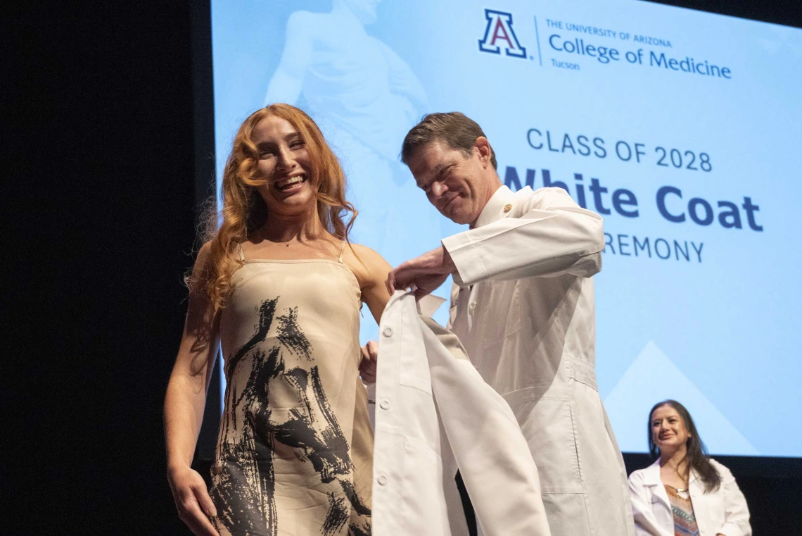 A University of Arizona College of Medicine – Tucson student in a tan print dress smiles as a professor helps her put on a white coat.