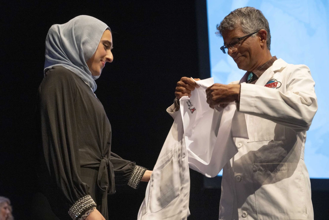 A University of Arizona College of Medicine – Tucson student wearing a black dress and gray hijab prepares to have a medical white coat put on her by a professor. 