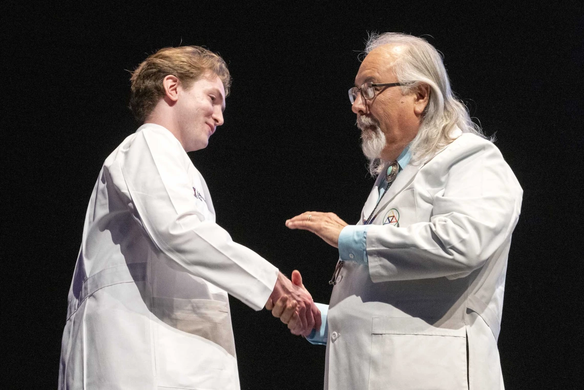 A University of Arizona College of Medicine – Tucson professor shakes hands with a new medical student. Both are wearing medical white coats.