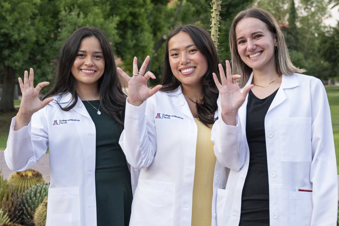 Three new University of Arizona College of Medicine – Tucson students wearing medical white coats smile as they flash the Arizona Wildcats hand sign.