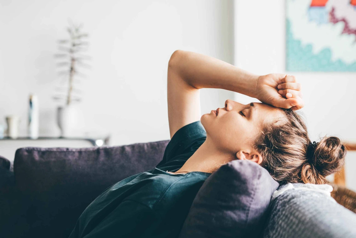 Person laying on a couch with arm raised and hand on forehead 