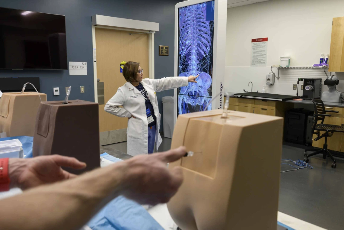 A professor in a white lab coat points to a projection of a skeleton while students in the foreground practice a procedure on the lower back of manikins. 