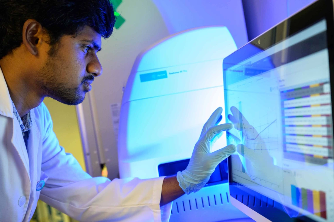 A graduate student in a white lab coat touches a computer monitor while looking at it intently. 