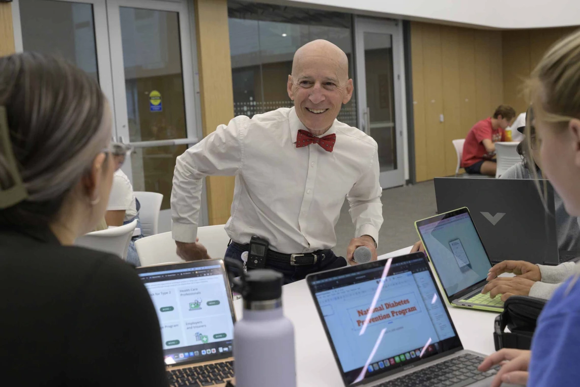 Dr. Paul Gordon smiles as he leans on a table in a classroom and talks with students who are on laptops. 