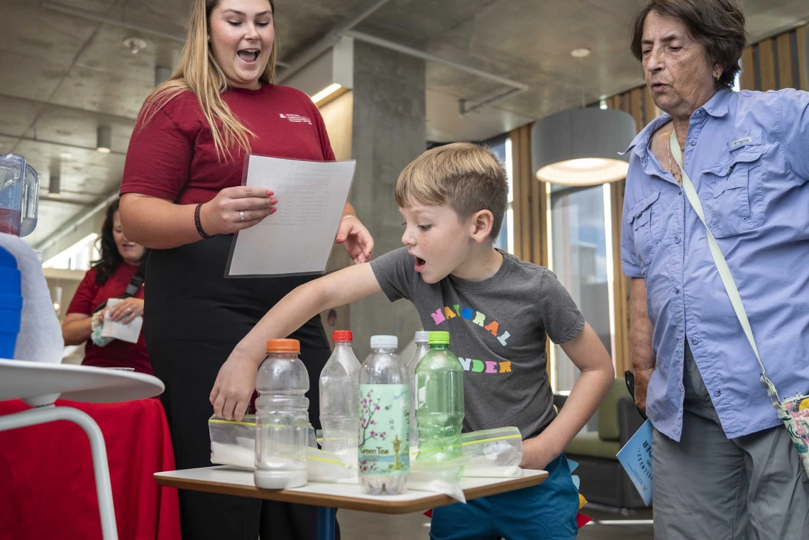 A surprised young boy lifts a bag of sugar behind soda bottles on a table while two women watch him. 