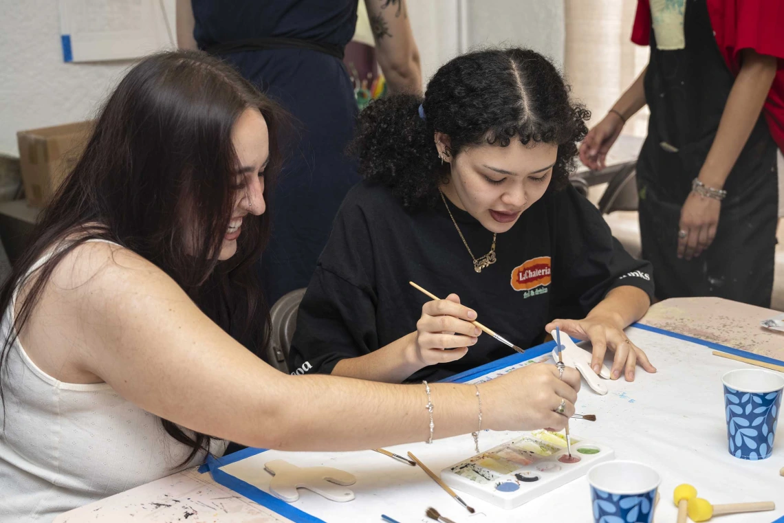 Two people sit at a table holding brushes as they paint tiles. 