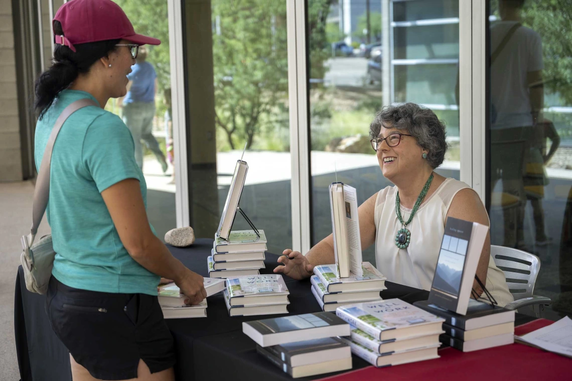 A smiling woman sits at a table covered by stacked books that she is signing while another woman stands at the table talking to her. 