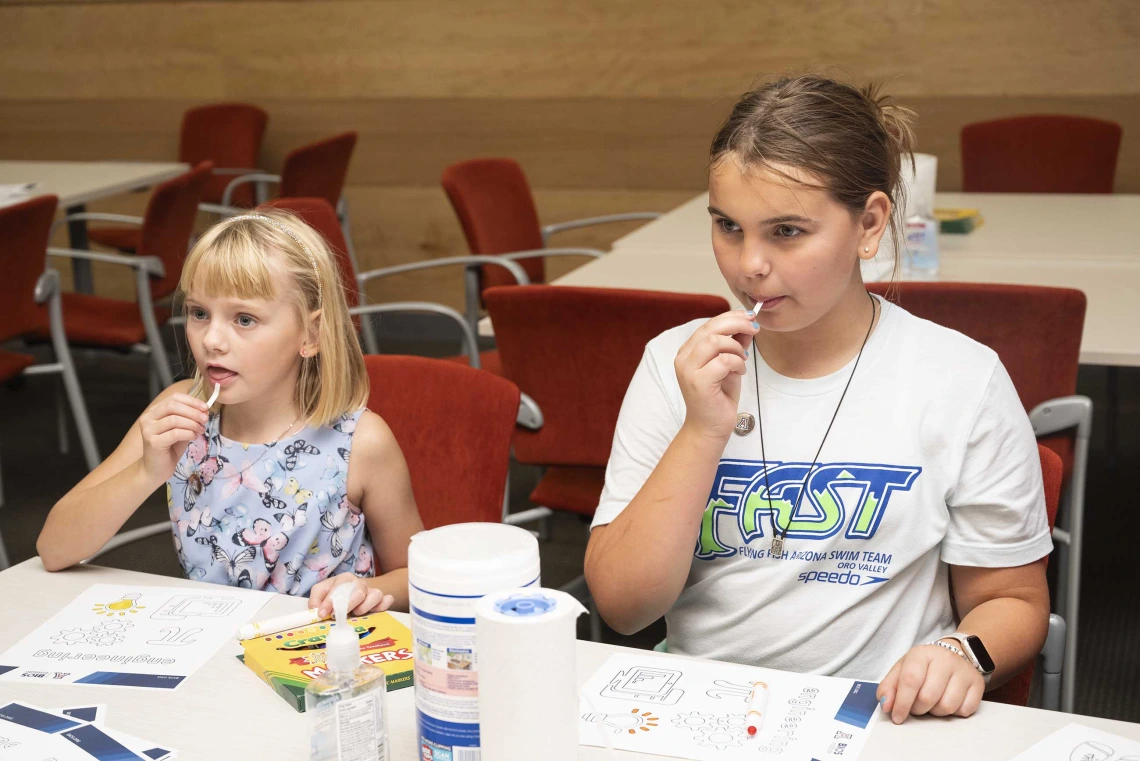 Two young girls sit at a table while they taste small pieces of flavored paper. 