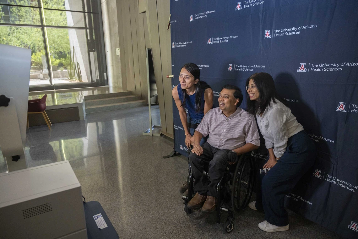 A man in a wheelchair is flanked by two women. All are smiling for a camera in front of a University of Arizona photo backdrop. 