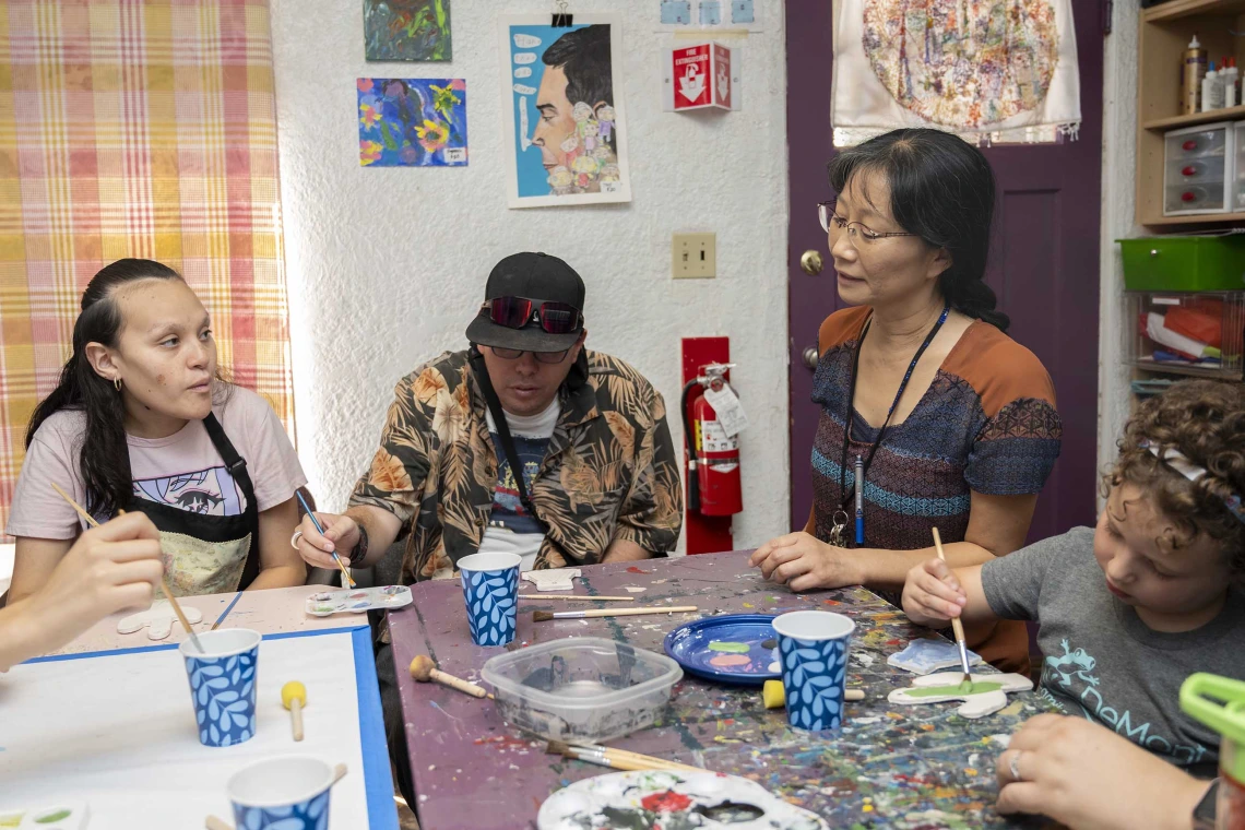 Yumi Shirai, PhD, sits at a table with a group of people who are painting at the University of Arizona’s ArtWorks program for adults with developmental disabilities. The table is covered with cups holding brushes and paints.