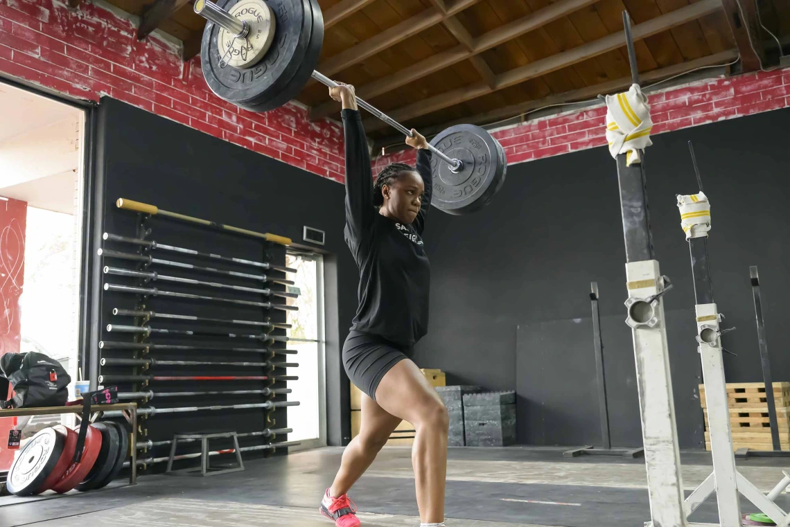 Photo of Brena Andrews in a gym holding a barbell over her head in split-jerk stance.