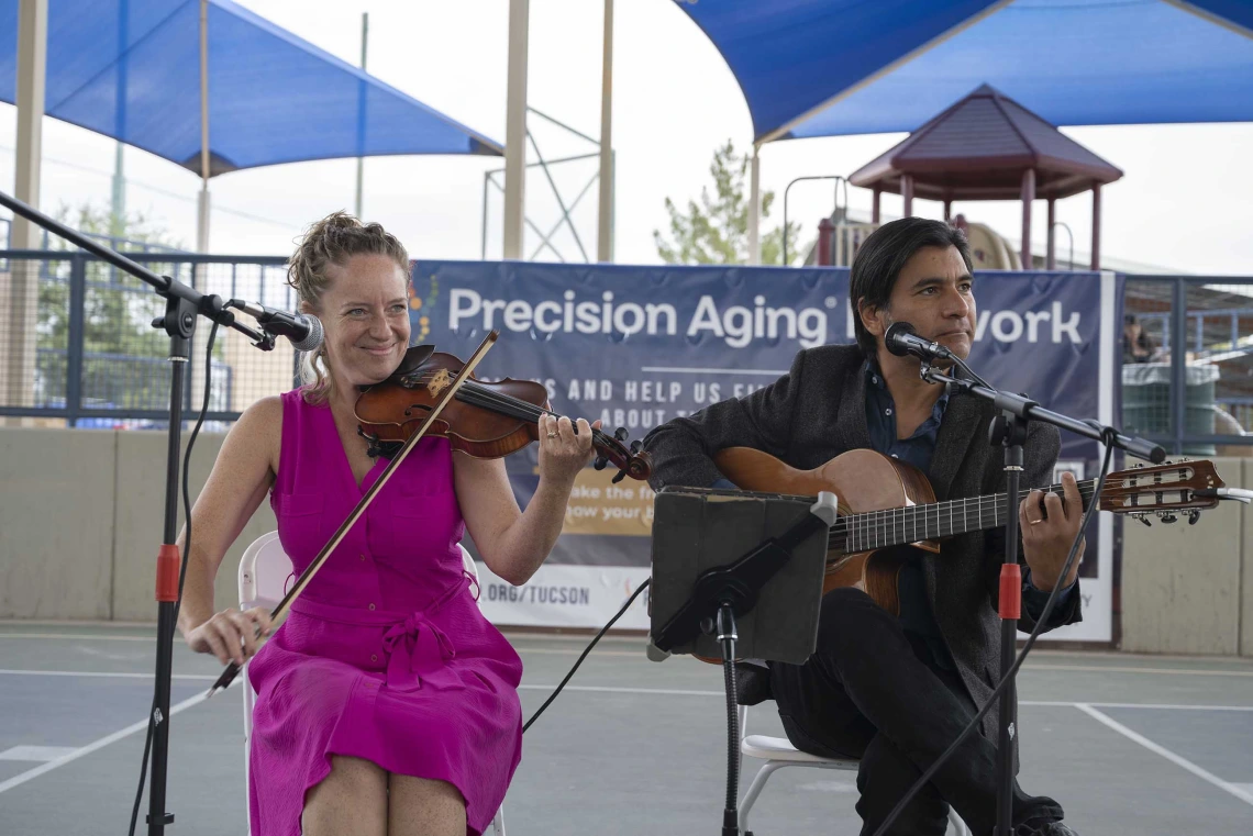 A seated woman plays a violin while a seated man plays a guitar in an outdoor settting. 