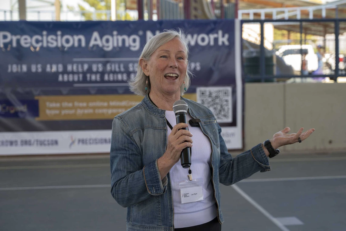 A woman stands on an outdoor sports court holding a microphone and speaking. She is smiling and holding up one hand as she speaks. 