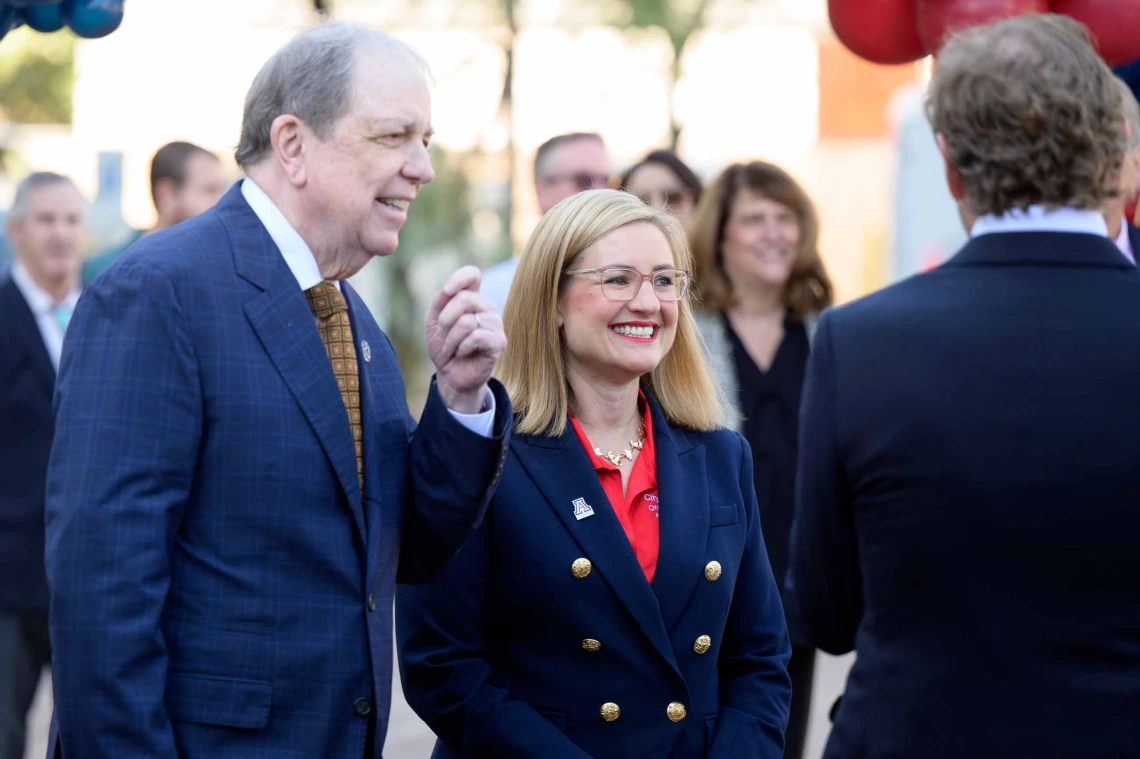 A man and woman dressed in dark blue suits stand and chat with others in an outdoor setting. Both are smiling. 