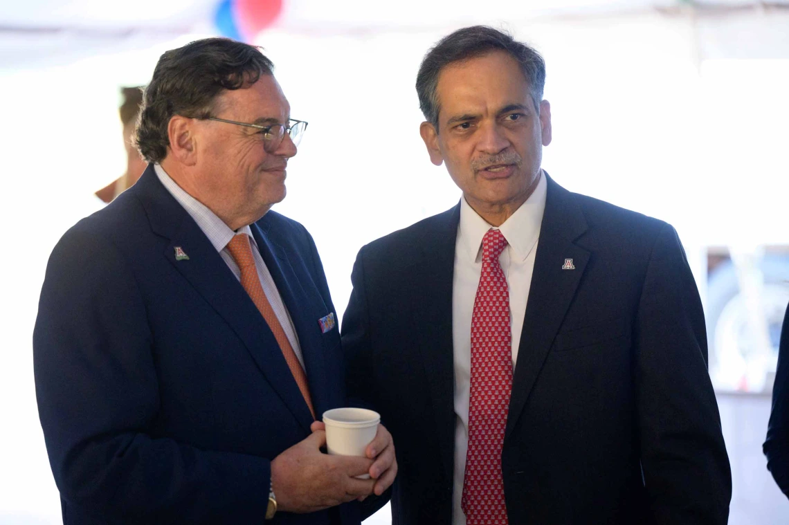 Two men in dark blue suits and red ties stand next to each other, chatting. 