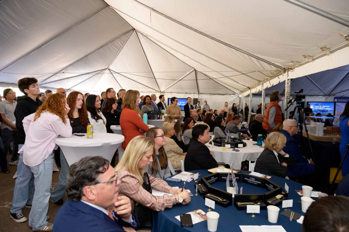 Hundreds of people sit and stand under a large white tent while listening to a speaker. 