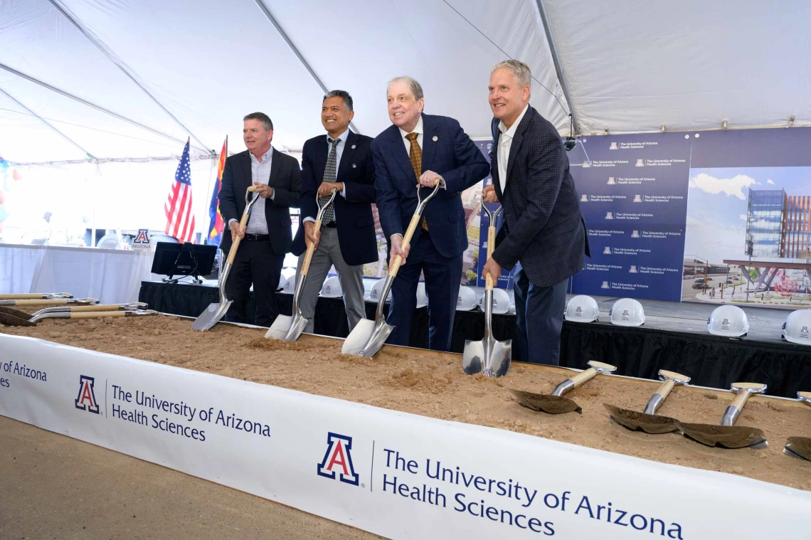 Four smiling men in suits hold shovels in sand with a University of Arizona Health Sciences banner in front of them.