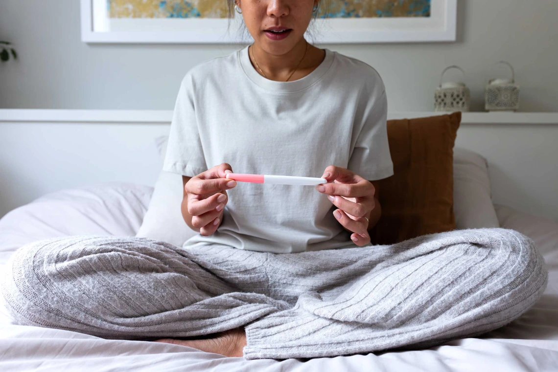 A young woman reads a pregnancy test as she sits up in bed wearing pajamas.