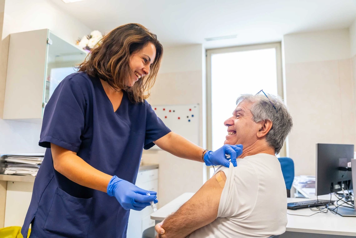 A female health worker chats with a seated male patient after administering a vaccine. Both are smiling.