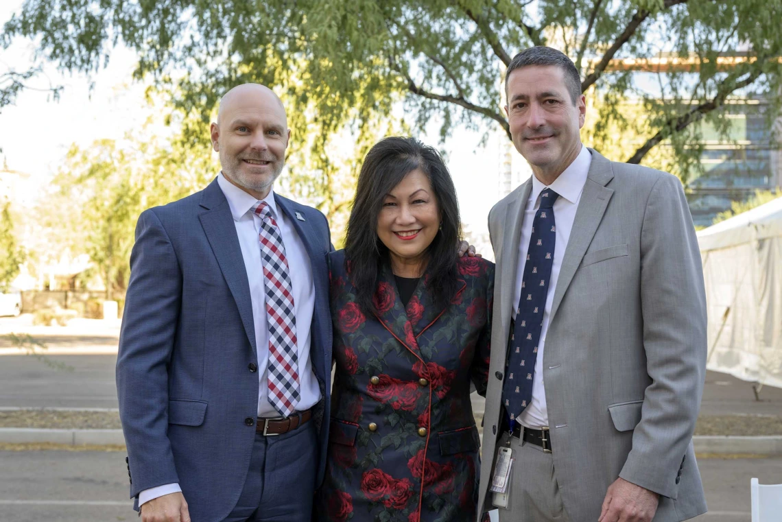 A woman flanked on either side by a man in a suit, stand outside under a tree. All three are smiling. 