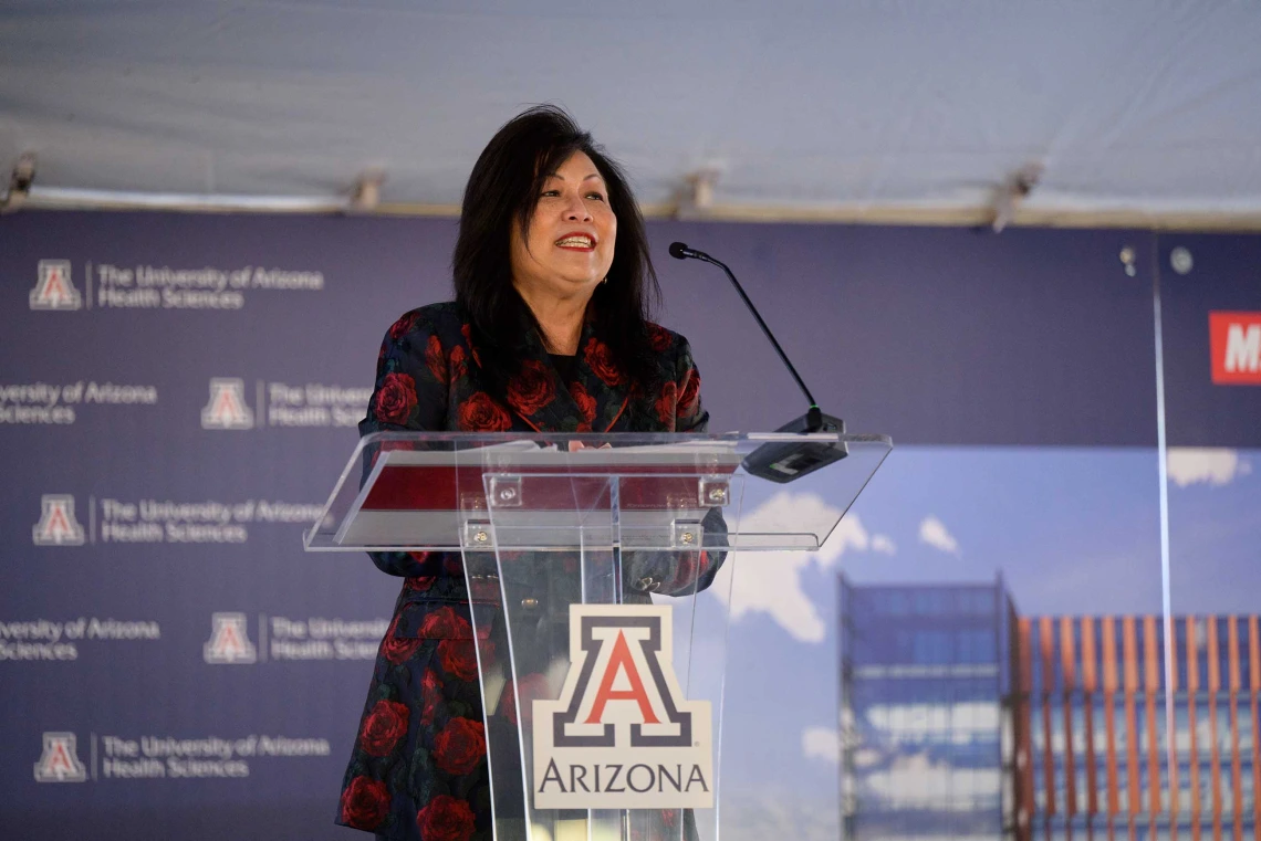 Cecilia Mata, chair of the Arizona Board of Regents, dressed in a dark blazer, stands at a lectern with the University of Arizona logo on it. 