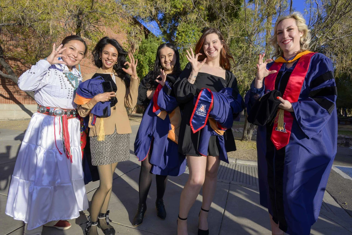 Five University of Arizona nursing students stand outside wearing graduation regalia as they smile and show the Arizona Wildcats hand sign. 