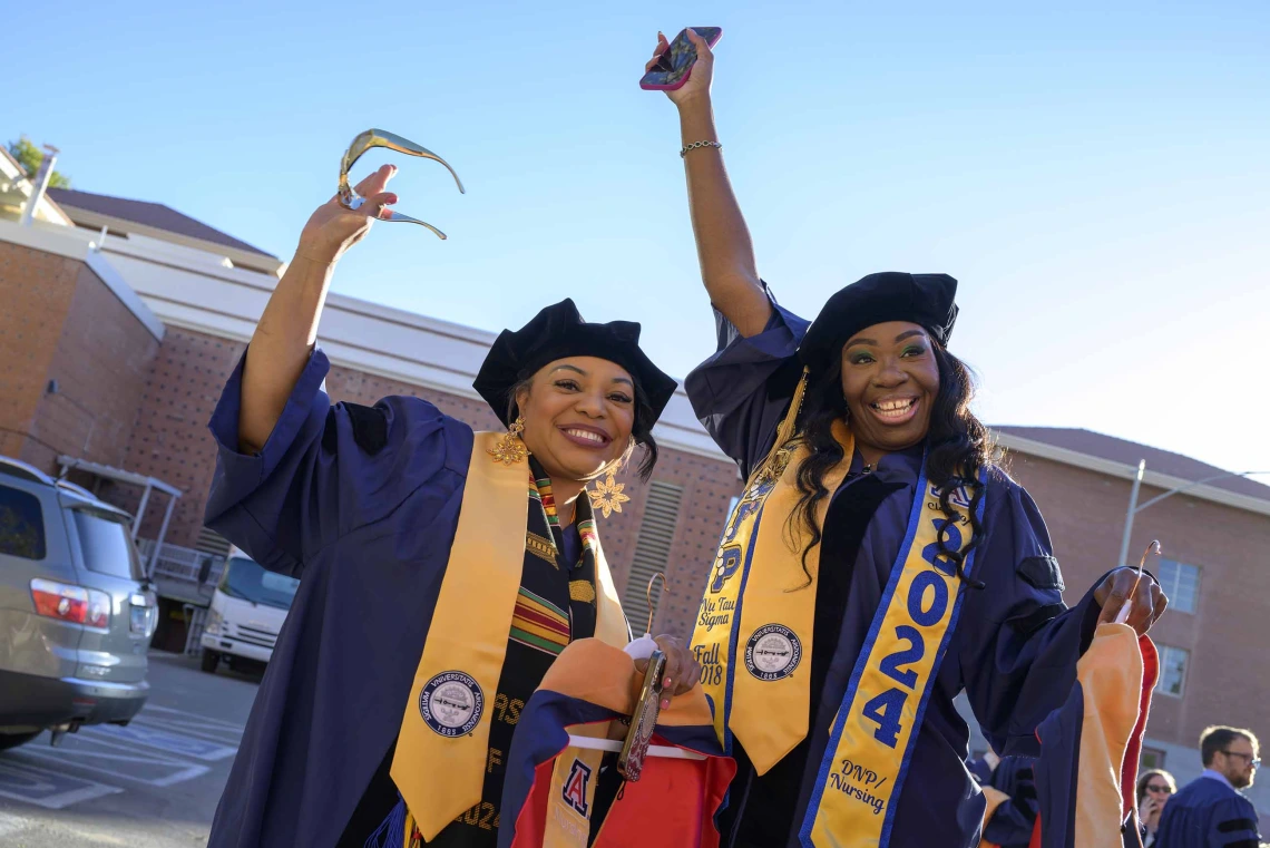 Two University of Arizona nursing students wave their arms in celebration while standing outside in their graduation caps and gowns. 