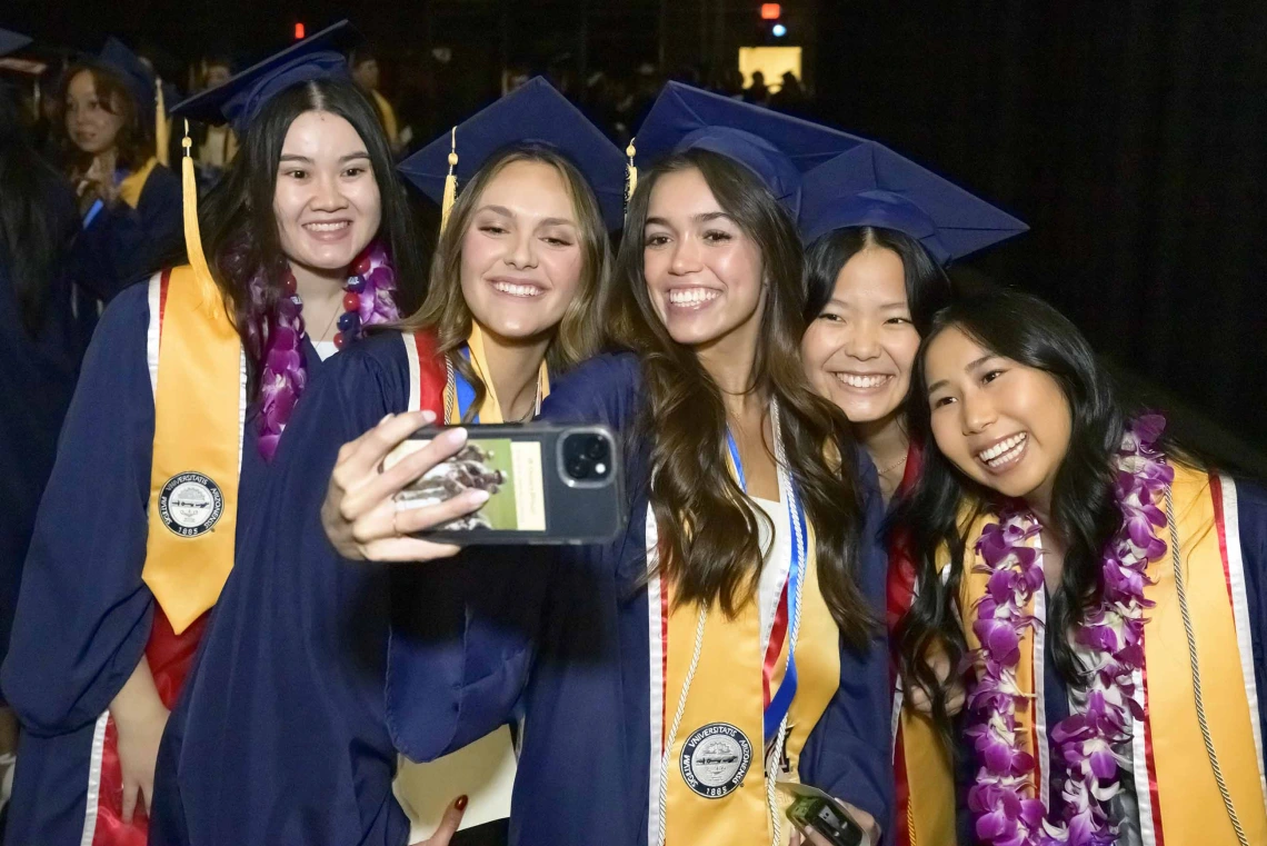 Five female nursing students wearing caps and gowns smile while taking a selfie as they stand side by side.