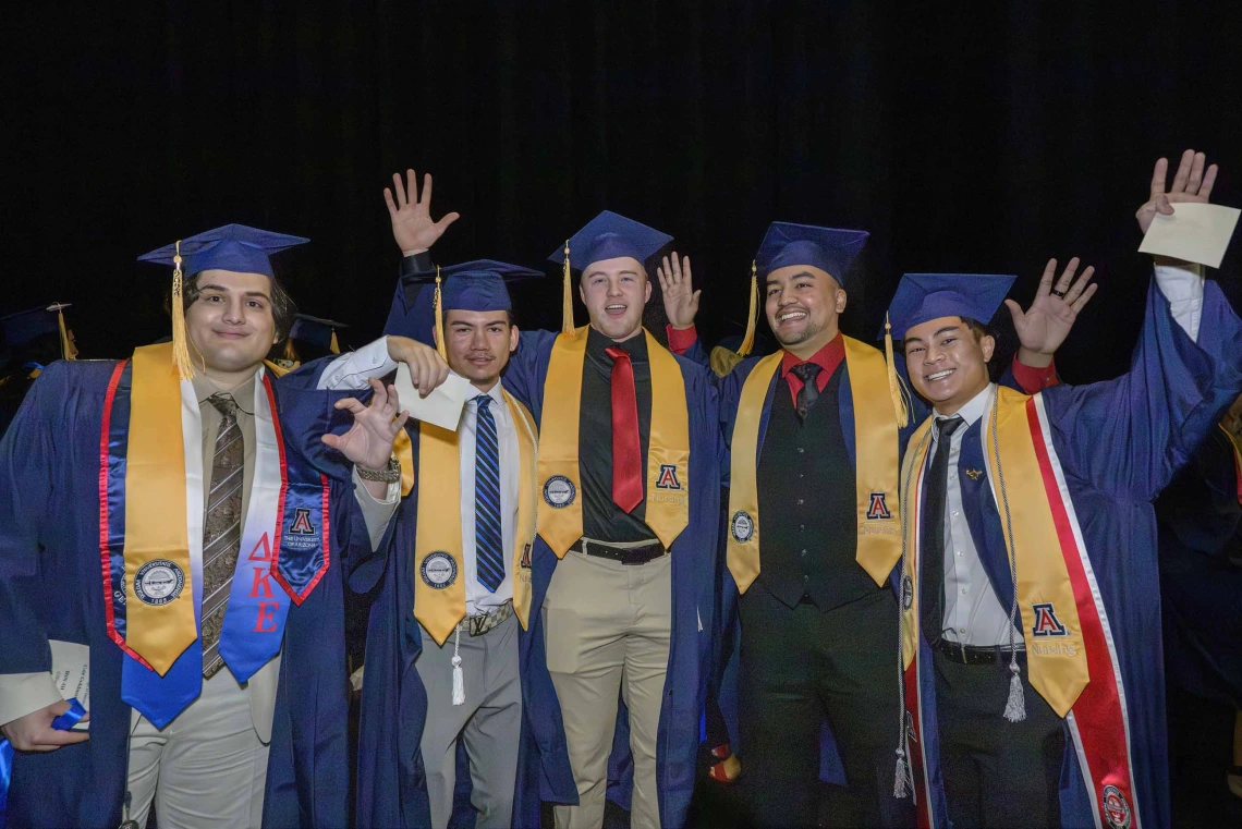 Five male nursing students wearing caps and gowns smile and wave as they stand side by side.