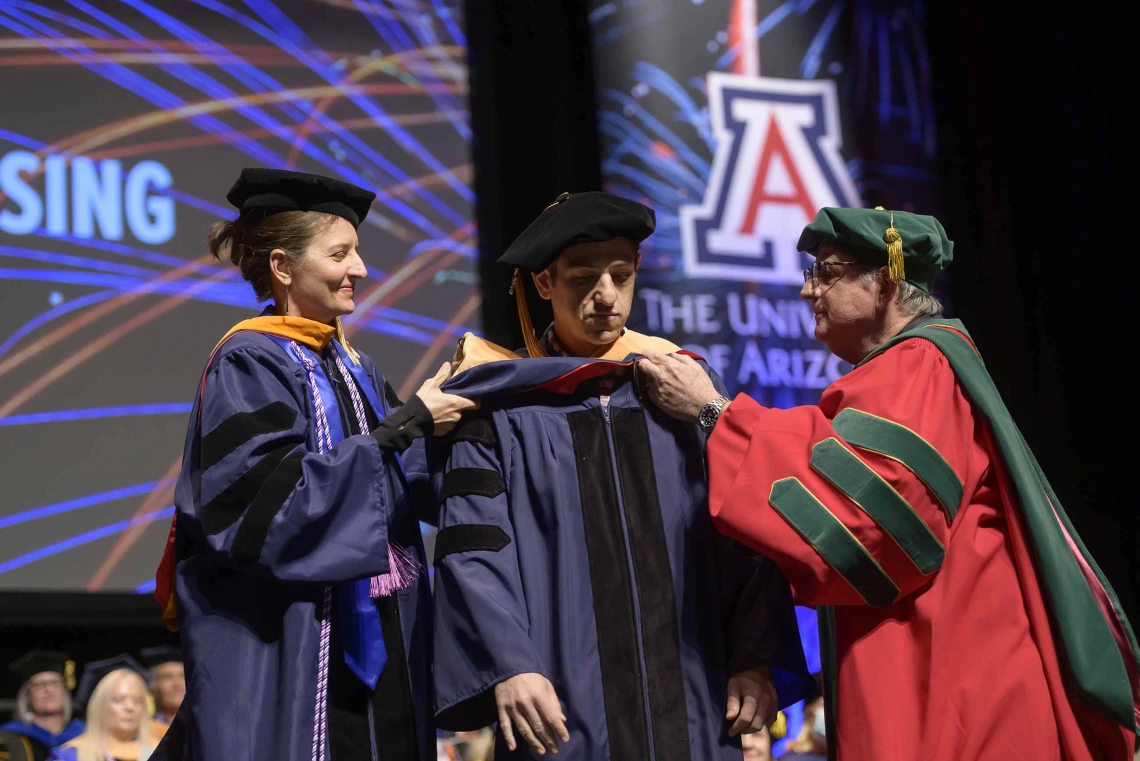 Two University of Arizona faculty members place a ceremonial hood over the shoulders of a student during a graduation ceremony. 