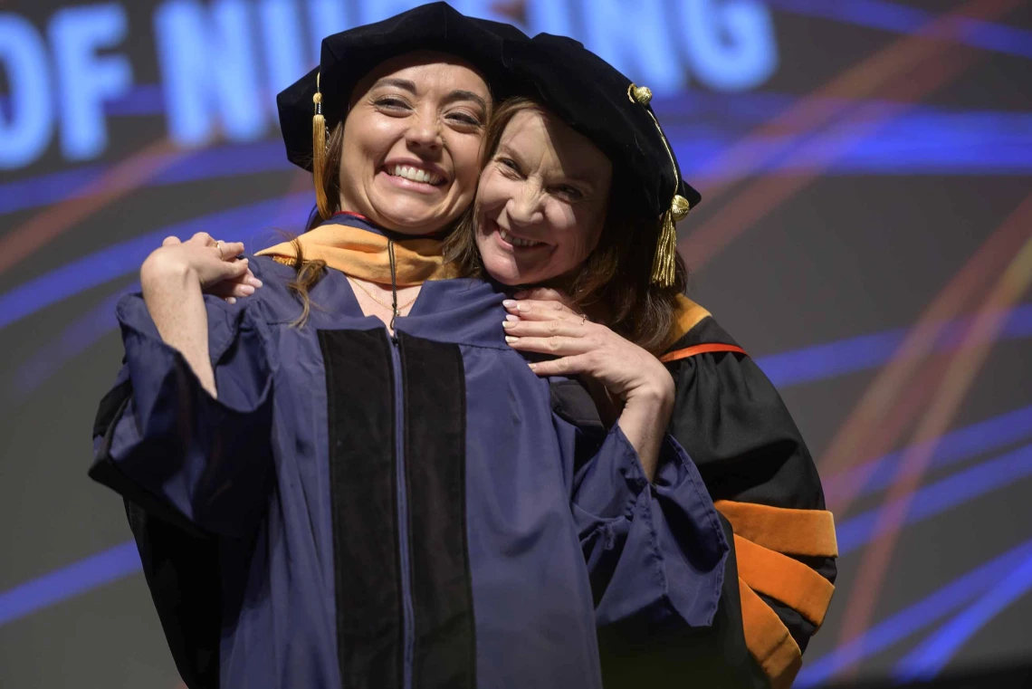 Two women in graduation regalia smile and hug. 