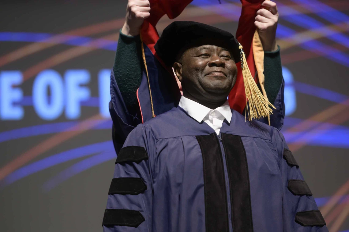 A Doctor of Nursing Practice student wearing a graduation cap and gown smiles as an unseen professor places a hood over his shoulders. 