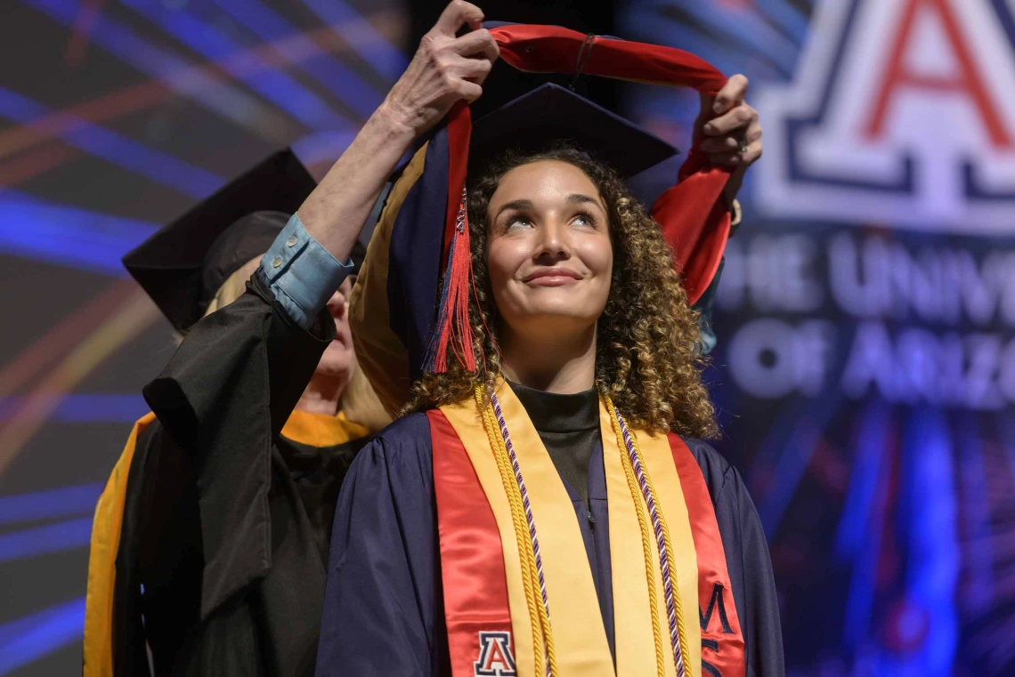 A woman wearing graduation regalia looks up and smiles as a traditional graduation hood is placed over her shoulders by a professor standing behind her. 