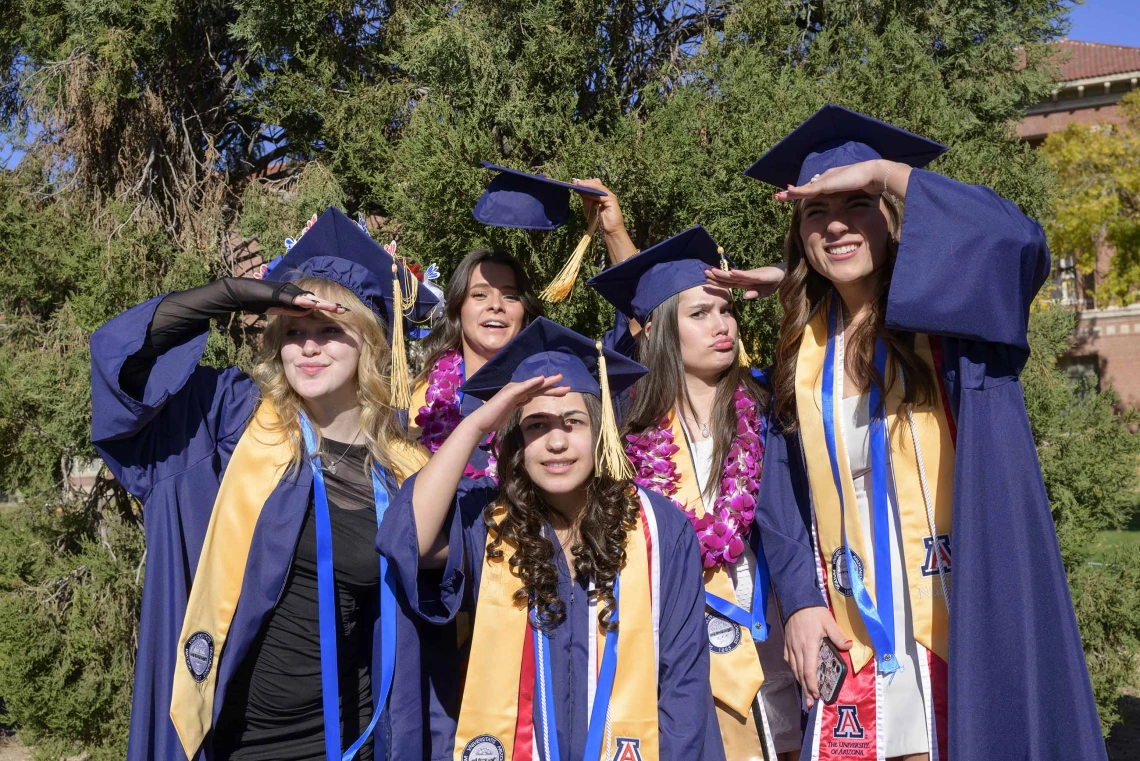 Five University of Arizona College of Nursing graduates in caps and gowns stand together, shielding their eyes from the sun. 