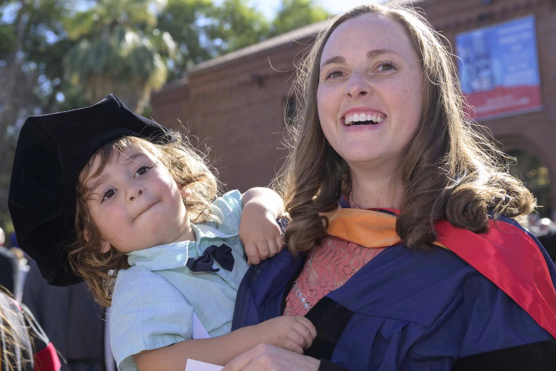 A smiling woman wearing a graduation gown holds a child who is wearing her graduation cap outside. 