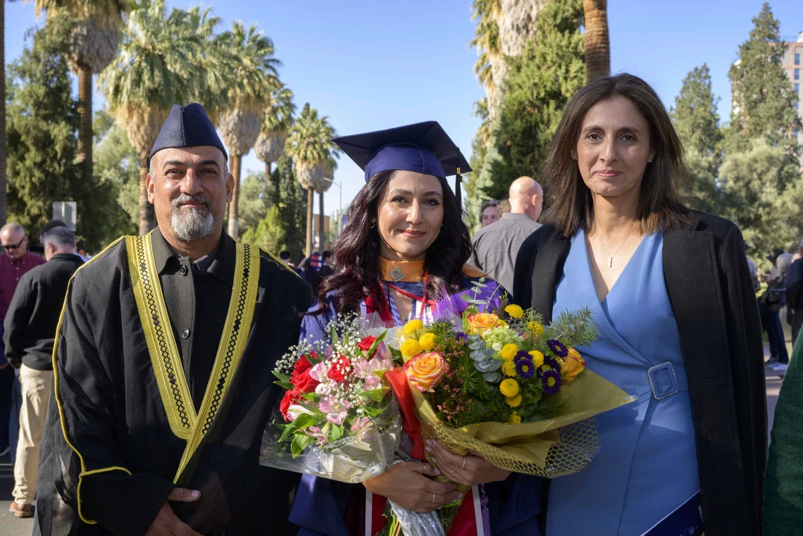 A young woman dressed in a graduation cap and gown holds flowers as she is flanked by her parents. 