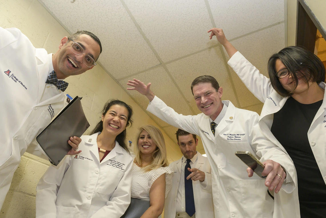 Six medical school faculty members wearing white coats laugh and make funny gestures with their arms while standing in a hall. 