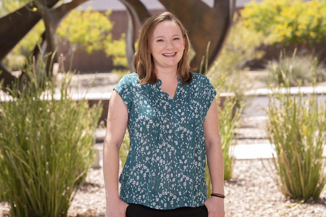 Woman smiling, wearing a green and black shirt, behind her are bushes and a tree.