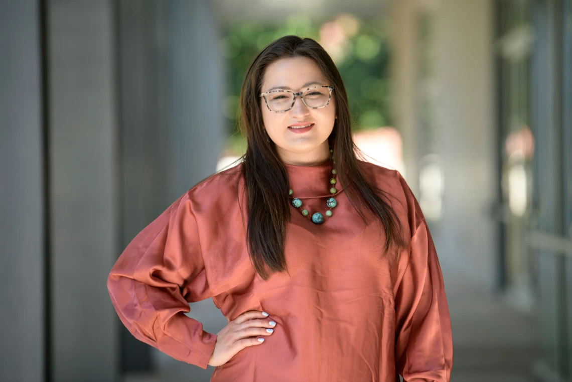 Woman with long dark hair and glasses wearing a bright red shirt and blue necklace stands confidently with a hand on her hip